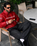 A man wearing a red Porsche leather jacket, black trousers, and sunglasses sits in a wooden chair at an outdoor cafe, holding a small espresso cup. The Porsche brand name is clearly visible on the chest of the jacket.
