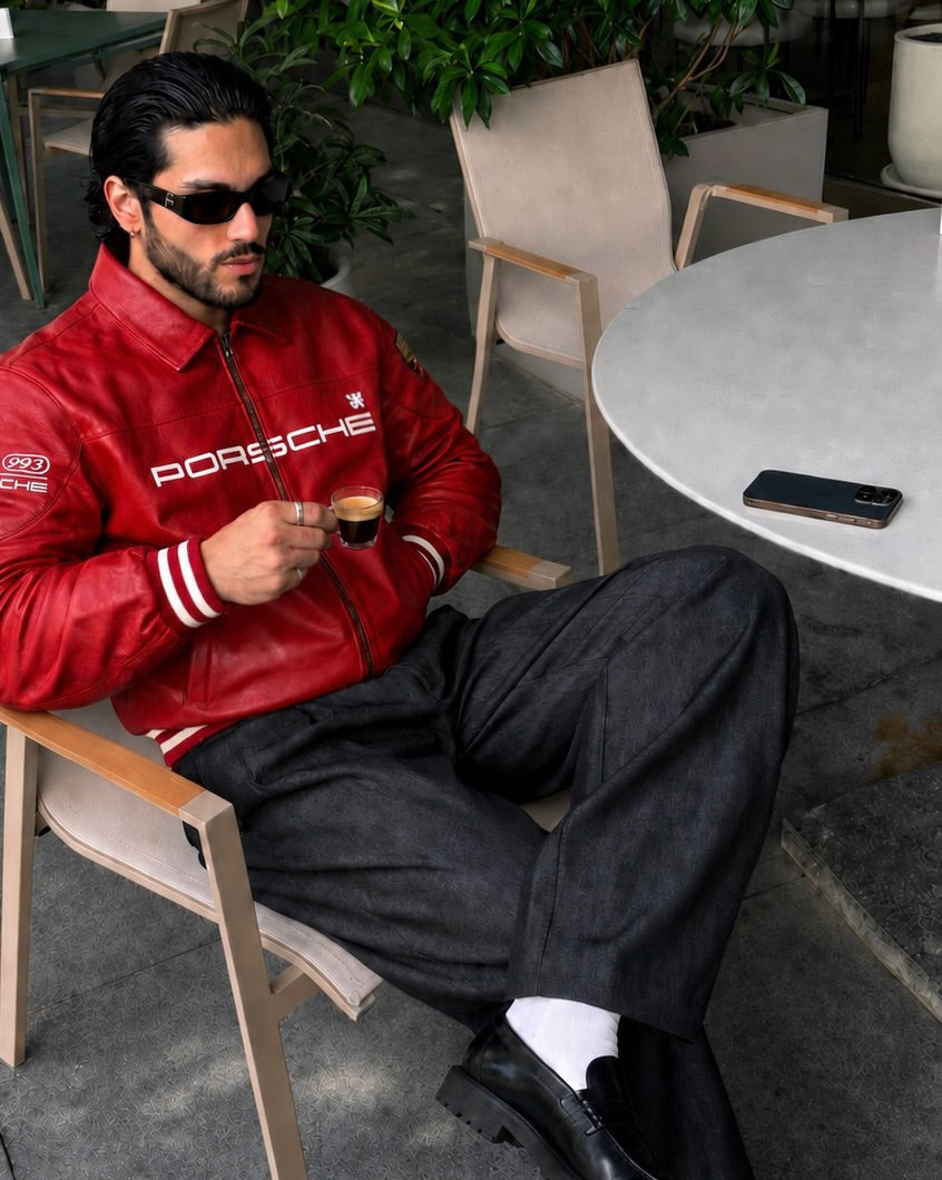 A man wearing a red Porsche leather jacket, black trousers, and sunglasses sits in a wooden chair at an outdoor cafe, holding a small espresso cup. The Porsche brand name is clearly visible on the chest of the jacket.