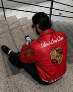 A man sitting on stone stairs wearing a red leather jacket with 'Amié Leon Dore' and a Porsche crest on the back, looking at his phone.